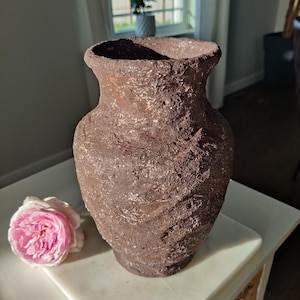 May include: A brown vase with a rough, textured surface and a wide, flared rim. The exterior features a rugged, uneven finish with subtle metallic highlights. A single pale pink rose with layered petals is positioned next to the base of the vase.