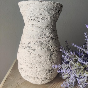 May include: A beige vase with a rough, textured surface and a wide, flared opening. The exterior features a mottled, uneven finish with dark grey undertones visible through the light-colored coating, creating a weathered and organic appearance.