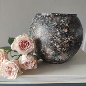 May include: A spherical vase with a textured black finish featuring distressed copper and silver highlights, accompanied by three pale pink roses. The vase has a rounded body and a wide opening, displaying a weathered appearance with visible brushstrokes.