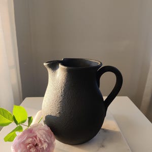 May include: A matte black ceramic pitcher with a textured, granular surface, a rounded body, and a curved handle. The pitcher features a distinct pouring spout at the rim. A single pale pink rose with green leaves is positioned next to the pitcher.