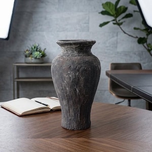 May include: A dark charcoal gray vase with a heavily textured, rugged surface. The vessel features a wide, flared rim that tapers down into a bulbous body, narrowing toward the base. The finish appears matte and weathered with deep vertical grooves.