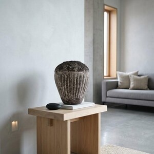 May include: A dark brown, textured stone vessel with vertical fluted carvings along its body, resting on a white rectangular base. Beside the vessel sits a small, smooth, black oval object on a light-colored wooden side table.