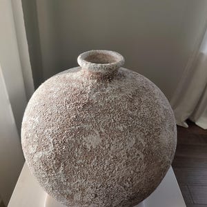 May include: A spherical vase with a textured, rough surface featuring a mottled blend of off-white and earthy brown tones. The vessel has a small, circular opening at the top with a slightly raised, rounded rim, showcasing a weathered appearance.