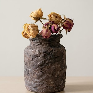 May include: A dark brown, cylindrical vase with a rough, textured surface containing a bouquet of dried roses in shades of pale yellow, deep pink, and muted red. The vase has a thick, uneven rim and a weathered, earthy appearance.