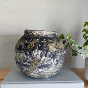 May include: A round, bulbous vase with a textured surface featuring a mottled blend of black, white, and metallic gold paint. The vessel has a wide, open mouth and a thick, uneven rim, showcasing a rugged, hand-crafted appearance with deep crevices.