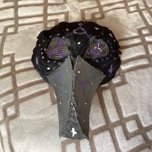 Peut inclure: Un masque en feutre noir avec un long bec pointu. Le masque a deux trous pour les yeux en maille violette et des détails peints en blanc et violet.