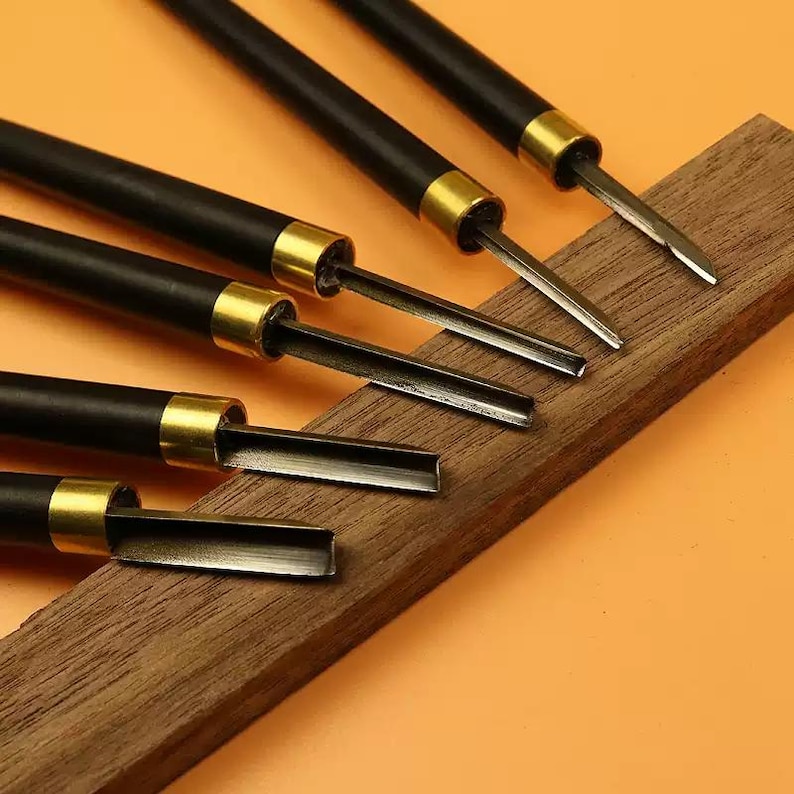 May include: Set of six wood carving tools with black handles and gold accents. The tools have different shaped blades and are arranged on a wooden surface.