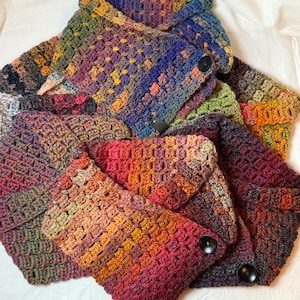 May include: A collection of colorful, crocheted cowls in various shades of red, orange, yellow, green, blue, and purple. Each cowl features a button closure and a small, rectangular tag. The handmade scarves are arranged on a white surface.