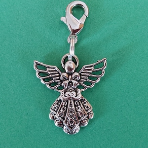 May include: Silver angel charm with a detailed design and a clip-on closure. The angel has wings, a bow, and a flowing dress.