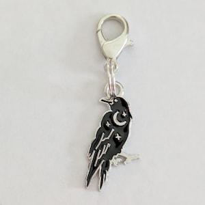May include: A silver-toned charm with a black raven design. The raven is perched on a branch and features a crescent moon and star accents. The charm is attached to a lobster clasp.