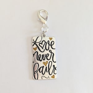 May include: A silver charm with the words "Love never fails" in black script, accented with gold hearts. The charm has a lobster clasp for easy attachment to a bracelet or necklace.
