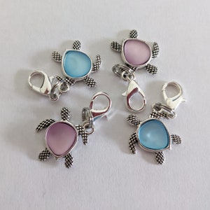 May include: Five silver-toned turtle charms with lobster claw clasps. Each turtle has a textured shell design and a colored oval center in either light blue or pink. The charms are on a white surface.