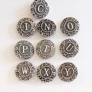 May include: Set of 10 silver-toned metal buttons with a decorative design and a letter from the alphabet in the center. The letters are C, I, N, O, P, T, U, W, X, and Y.