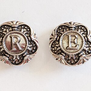 May include: Two silver-toned metal buttons with ornate designs. The buttons feature the letters "R" and "E" in the center.