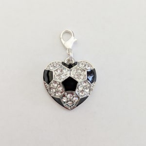 May include: A heart-shaped charm with a soccer ball design. The charm features a silver clasp and is adorned with clear rhinestones and black enamel. The soccer ball pattern is visible on the front of the charm.