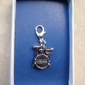May include: A silver-colored charm in the shape of a drum set, including a bass drum and two tom-toms. The charm is attached to a silver clasp. The charm is displayed inside a blue box with a white border.