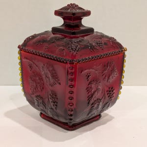 May include: A ruby red glass candy dish with a square base and lid. The dish features embossed grape and leaf designs. The lid has a square handle. The edges are trimmed with small, yellow-tinted beads.