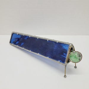 May include: A handmade kaleidoscope with a rectangular blue stained glass body and a silver metal frame. The kaleidoscope has a small green glass lens at one end and rests on three small metal legs.