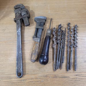 May include: A collection of vintage hand tools, including a large pipe wrench, adjustable wrench, a screwdriver, and several drill bits. The tools are arranged on a light brown wooden surface.