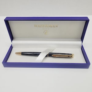 May include: A black Waterman pen with gold-colored accents rests inside a blue presentation box. The box is lined with white fabric and features the Waterman logo in gold lettering.