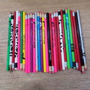 Puede incluir: Una colección de lápices de varios colores, incluyendo verde, rosa y rojo, con textos como "BEEF gives you zip!" y "ZINC - IRON - PROTEIN." Algunos lápices tienen diseños de vaca. Los lápices están dispuestos sobre una superficie marrón claro.