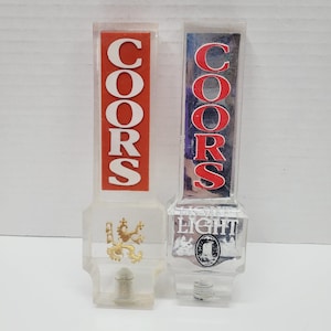 May include: Two clear plastic Coors beer tap handles. One has a red background with white lettering, the other has a silver background with red lettering. One has a gold emblem, the other says "LIGHT".