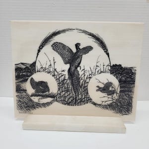 May include: A rectangular, off-white decorative plaque featuring a black and white illustration of a pheasant in a field. The artwork includes wheat stalks and two smaller circular images of pheasants. The plaque is supported by a matching stand.