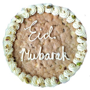 May include: A round cookie with a light brown base, decorated with white frosting swirls around the edge. The cookie is inscribed with the words "Eid Mubarak" in white icing. Gold sprinkles and pistachio pieces add decorative accents.
