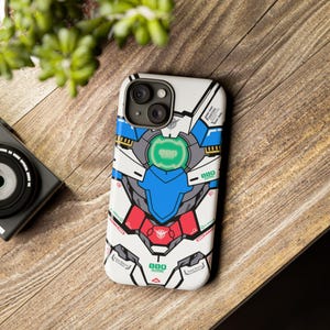 May include: White phone case with a colorful graphic design. The design features a robot-like illustration with blue, red, and green accents. The case is on a wooden surface, with a camera and greenery in the background.