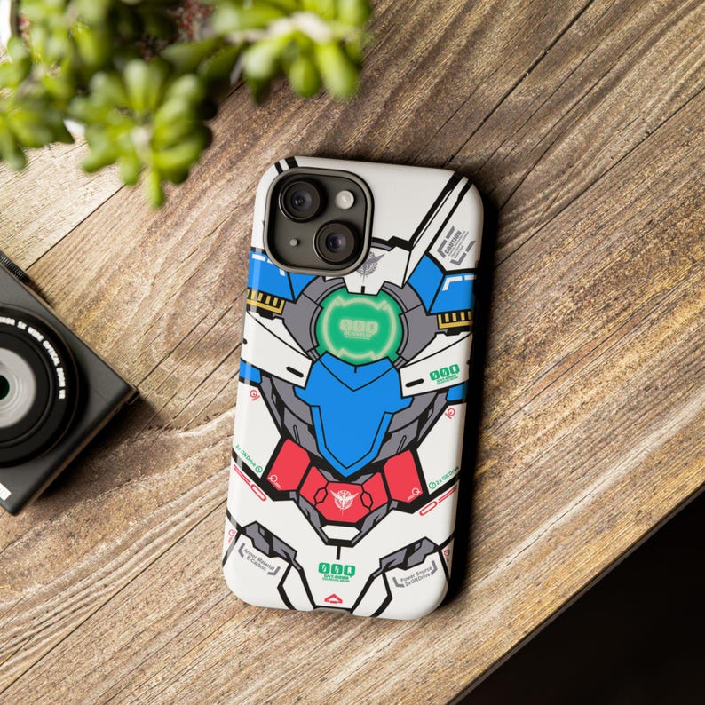 May include: White phone case with a colorful graphic design. The design features a robot-like illustration with blue, red, and green accents. The case is on a wooden surface, with a camera and greenery in the background.