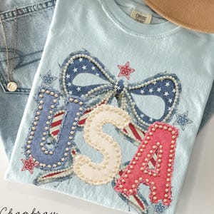 May include: Light blue t-shirt with a patriotic "USA" design. The letters are embellished with pearls and red, white, and blue accents. A brown hat and blue jeans are also in the image. The word "Chambray" is written in cursive at the bottom.