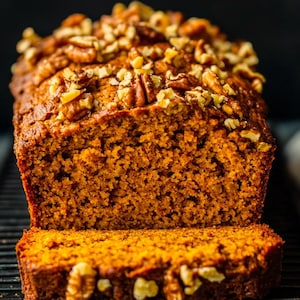 May include: A loaf of pumpkin bread with a slice cut out, resting on a black cooling rack. The bread is a warm orange color and topped with chopped walnuts. The background is dark, highlighting the bread's texture and details.