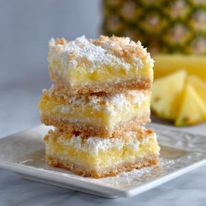 May include: A stack of three square dessert bars on a white plate. The bars have a golden-brown crust, a yellow filling, and a white, shredded coconut topping. Slices of pineapple are in the background.