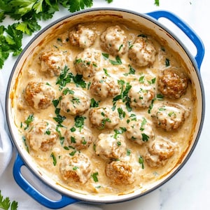 Puede incluir: Una cazuela de cerámica blanca con un asa azul llena de albóndigas suecas en una salsa cremosa y adornada con perejil fresco.