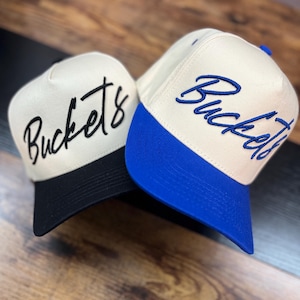 May include: Two baseball caps with the word "Buckets" embroidered on the front. One cap has a black brim and black lettering, while the other has a blue brim and blue lettering. The caps have a cream-colored crown.