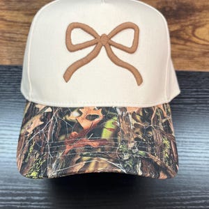 May include: A beige baseball cap with a brown bow design on the front. The brim features a camouflage pattern in shades of brown, green, and orange. The cap is made of a sturdy material and has a classic, structured shape.