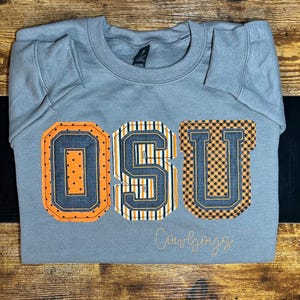 以下が含まれることがあります： グレーのトレーナーに、オレンジ、黒、白で「OSU Cowboys」という大きな刺繍が施されています。文字は大学風のフォントです。