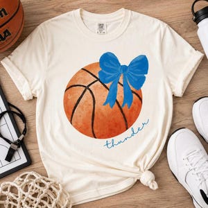 Op de afbeelding: Crèmekleurig T-shirt met een aquarel basketbalontwerp en een blauwe strik. Het woord "thunder" staat in blauwe letters eronder. Het shirt wordt gestyled met witte sneakers en een waterfles.