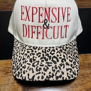 Könnte beinhalten: Eine weiße Baseballkappe mit den rot gestickten Worten "EXPENSIVE & DIFFICULT". Der Schirm hat ein Leopardenmuster in Schwarz und Beige. Ein modisches Accessoire.