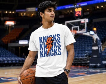 Op de afbeelding: Een wit T-shirt met korte mouwen waarop het woord KNICKS drie keer verticaal onder elkaar in het blauw is gedrukt, met een oranje bliksemschicht in de vorm van een basketbal in het midden van het tekstontwerp.