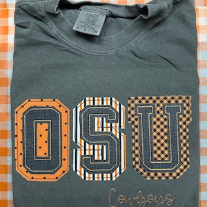 May include: Dark gray t-shirt with the letters "OSU" in orange, black, and white. The letter "U" has a black and orange plaid pattern. The word "Cowboys" is embroidered in a cursive font.