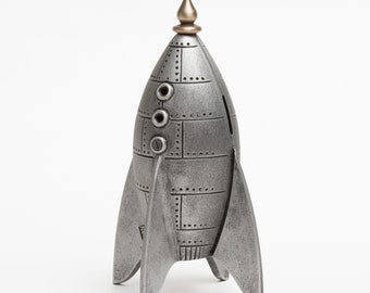 May include: A silver rocket-shaped money box with a gold finial on top. The money box has a slot on the back for coins.