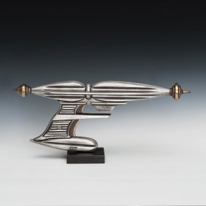 May include: A silver-colored futuristic-style ray gun with a brass-colored tip and handle. The gun is on a black stand with the word "Nelken" printed on it.