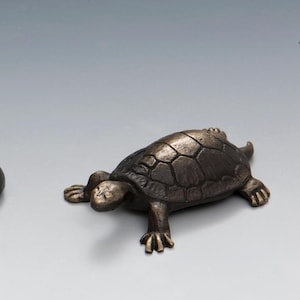 May include: A bronze turtle figurine with a detailed shell and a realistic pose. The turtle is facing to the right and has its head slightly raised.