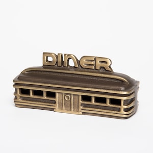 May include: A gold-colored metal model of a diner with the word "DINER" in raised letters on the roof. The model has a brown roof and gold-colored walls with windows.