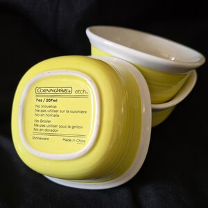 May include: Two square stoneware bowls with a bright yellow exterior and a white interior. The base of the bowl features the text CorningWare etch, 7oz / 207ml, No Stovetop, No Broiler, Stoneware, and Made in China, along with French and Spanish translations.