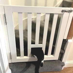 May include: White wooden pet gate with a black and white cat peeking through the bottom. The gate is installed at the top of a carpeted staircase.