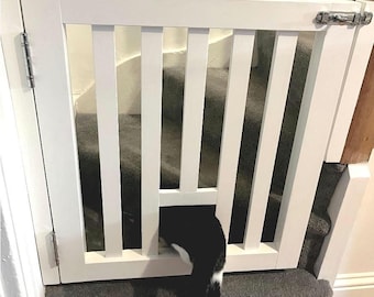 May include: White wooden pet gate with a black and white cat peeking through the bottom. The gate is installed at the top of a carpeted staircase.