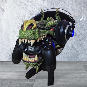 以下が含まれることがあります： 緑と金のモンスターをテーマにしたゲーム用ヘッドセットとコントローラーのスタンド。スタンドは、鋭い歯を持つ詳細なモンスターの頭部が特徴で、黒いPlayStationコントローラーを持っています。黒いヘッドセットがスタンドの背面に置かれています。