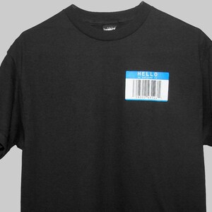 Puede incluir: Una camiseta negra con una etiqueta azul que dice "HELLO" y tiene un código de barras que dice "28002 69144".